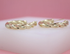 Two glossy, twisted gold hoop earrings rest side-by-side on a white surface against a soft pink background.