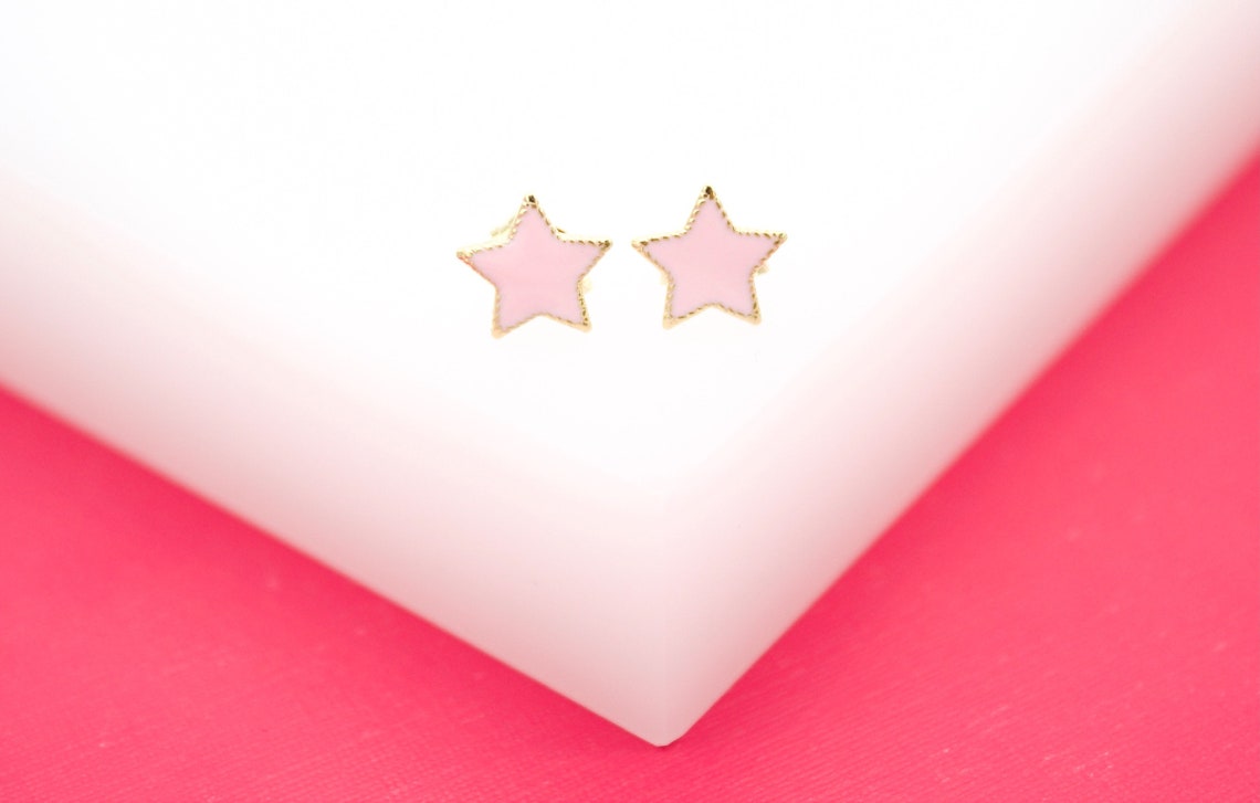 A pair of gold star stud earrings with light pink enamel details rests on a white and pink surface.