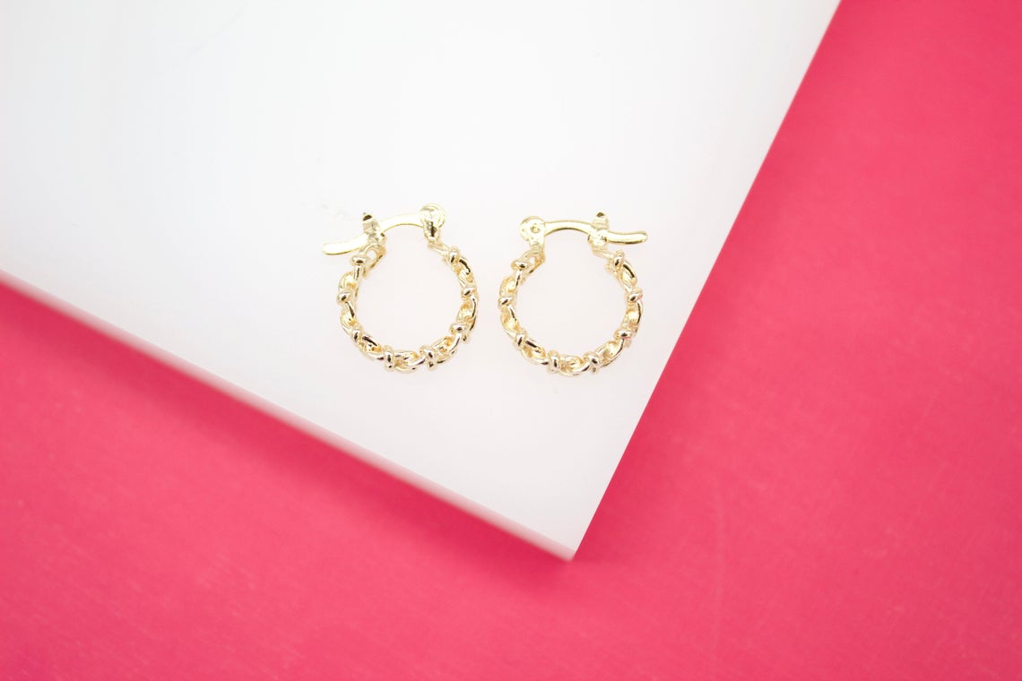 A pair of gold textured hoop earrings rests on a white display surface against a vibrant pink background.