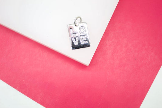 A silver rectangular pendant charm featuring the word "LOVE" spelled out in sparkling cubic zirconia stones rests on a white display against a vibrant pink background.
