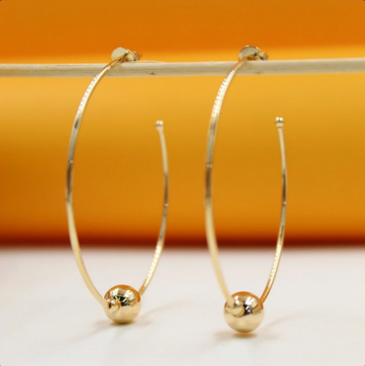 A pair of sleek gold hoop earrings featuring dangling round beads hangs delicately from a wooden stick against a warm orange and white background.