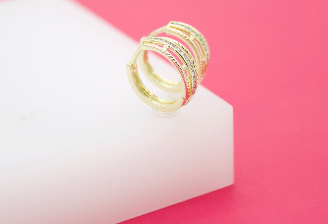 A pair of gold huggie hoop earrings featuring sparkling pavé crystals rest on a white display block against a vibrant pink background.