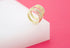 A pair of gold huggie hoop earrings featuring sparkling pavé crystals rest on a white display block against a vibrant pink background.