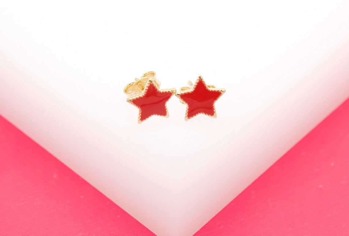 A pair of gold star stud earrings with vibrant red enamel centers rest on a clean white surface against a bright pink background.