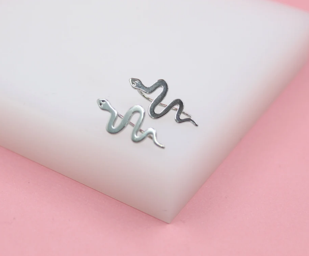 A pair of silver snake stud earrings rests on a white display block against a soft pink background.