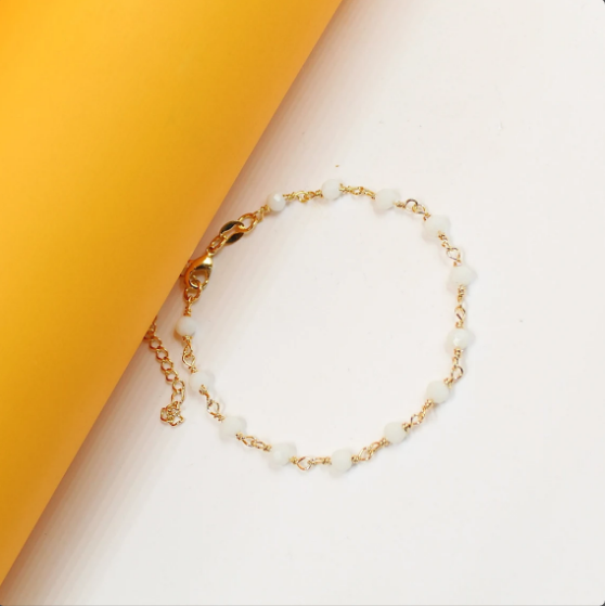 A delicate gold chain bracelet features small, translucent white beads arranged on a white and yellow background.