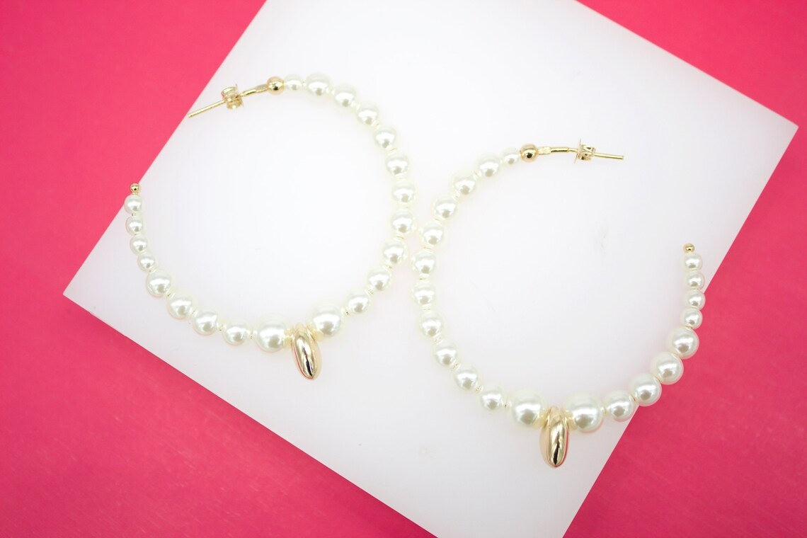 A pair of gold hoop earrings embellished with white pearls and featuring a central gold charm rests on a white display surface against a vibrant pink background.