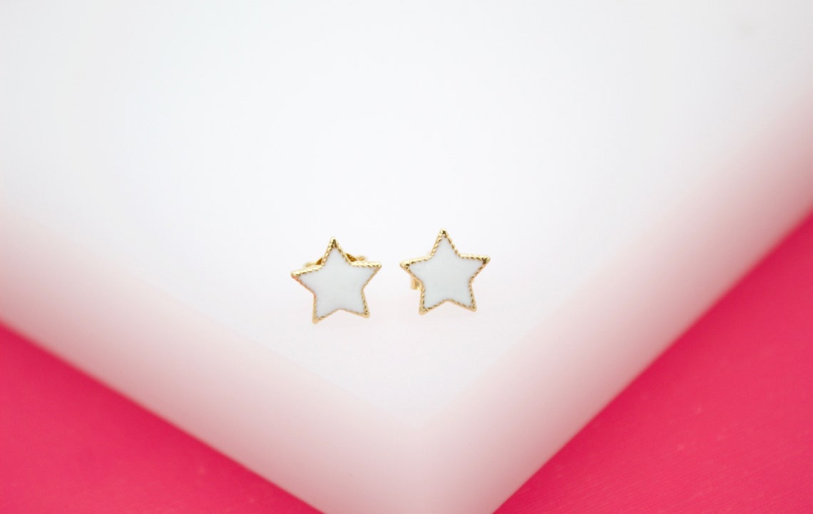 A pair of gold star stud earrings with white enamel fill rests on a white display against a pink background.
