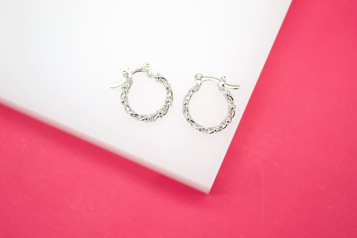 A pair of shiny silver textured hoop earrings rest on a white display against a vibrant pink background.