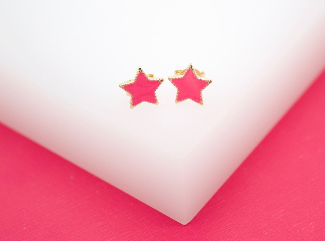 A pair of gold star stud earrings with bright pink enamel rests on a white display block against a vibrant pink background.