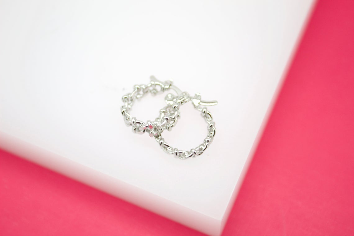 A pair of polished silver chain-link hoop earrings rests on a white display block against a vibrant pink background.
