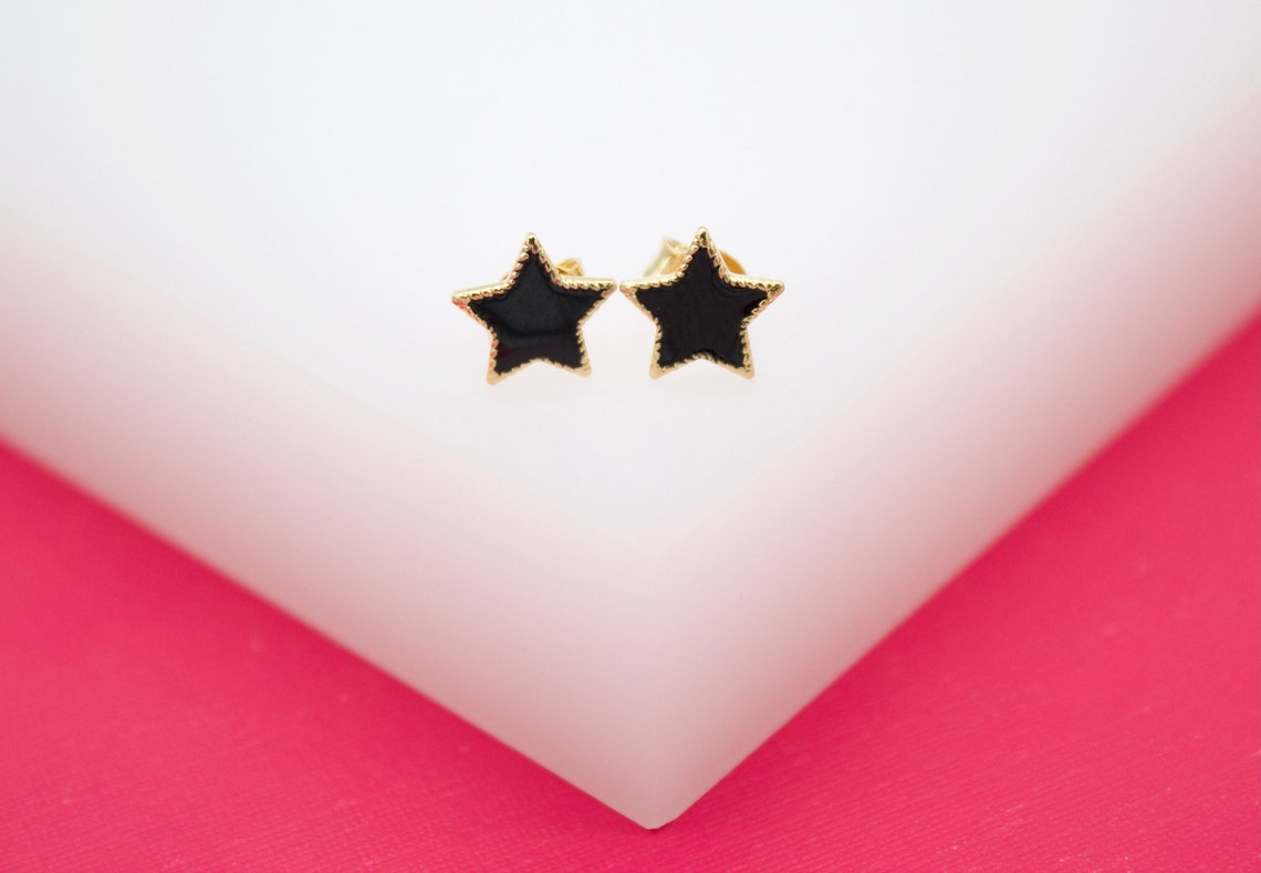 A pair of gold star stud earrings with black enamel display against a white and pink background.
