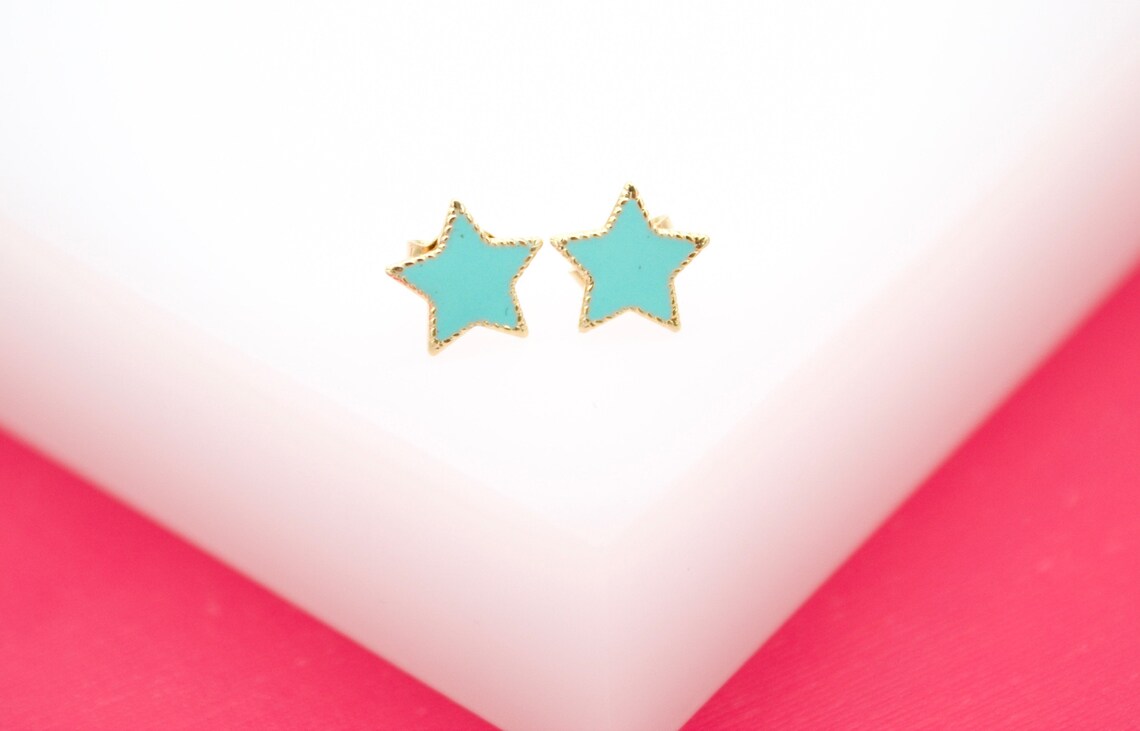 A pair of gold star stud earrings featuring vibrant turquoise enamel rests on a white display block against a bright pink background.