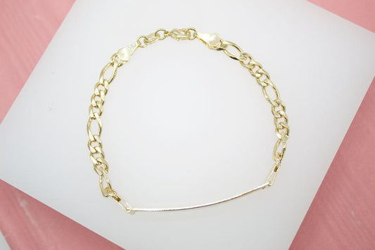A gold Figaro chain bracelet with an ID bar rests on a white display surface against a pink background.
