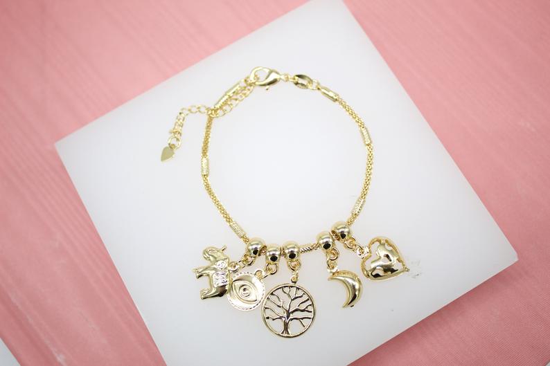 A gold charm bracelet featuring dangling symbols like an elephant, evil eye, tree of life, crescent moon, and heart rests on a white display against a pink background.