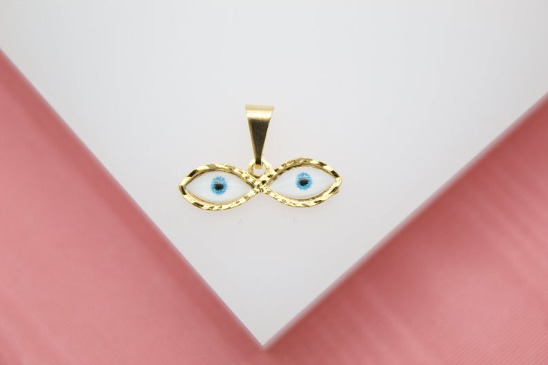 A gold evil eye pendant rests on a white and pink display surface, showcasing detailed blue and white enamel eyes.