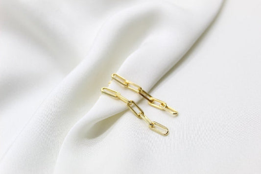 A pair of elegant gold paperclip chain earrings rests on a soft white silk fabric.