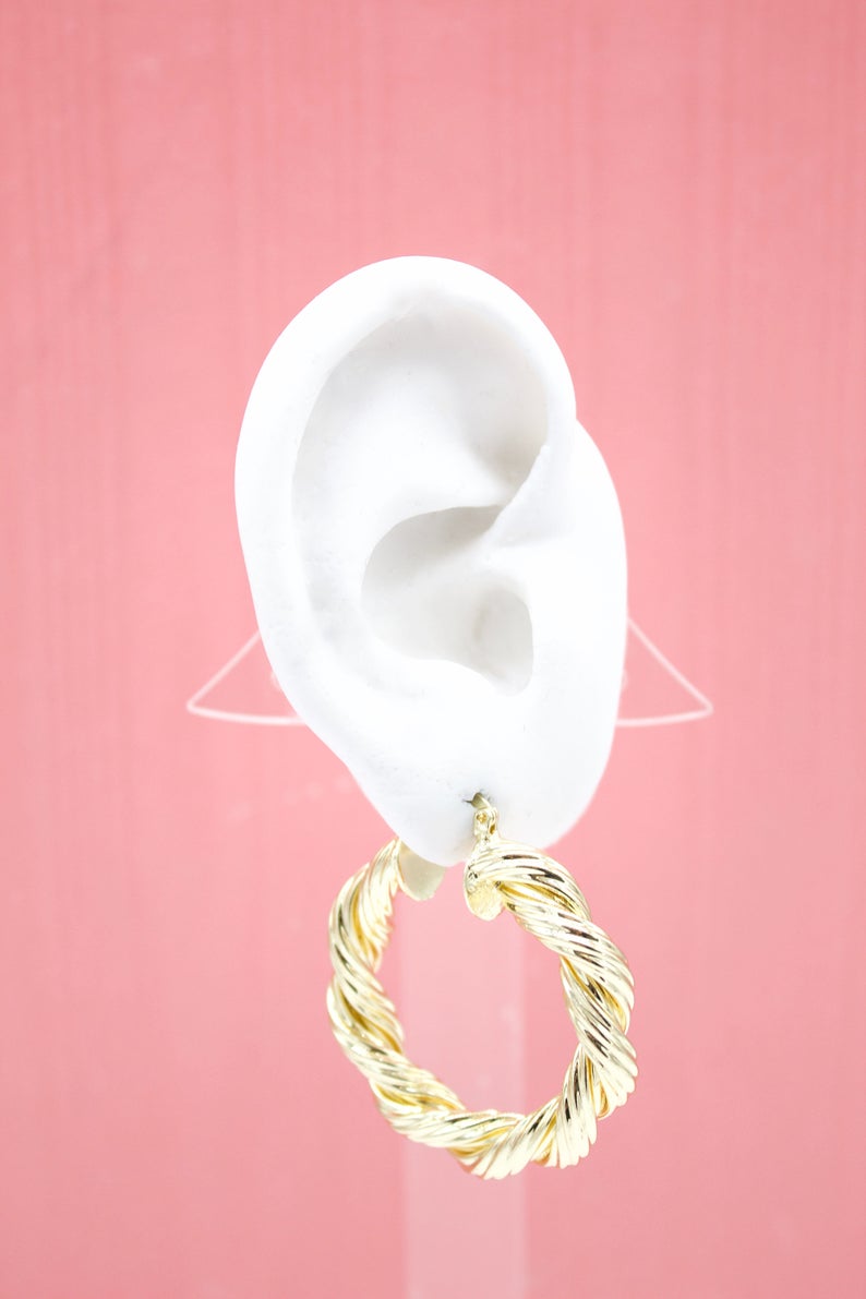 A white mannequin ear displays a chunky, twisted gold hoop earring against a solid pink background.
