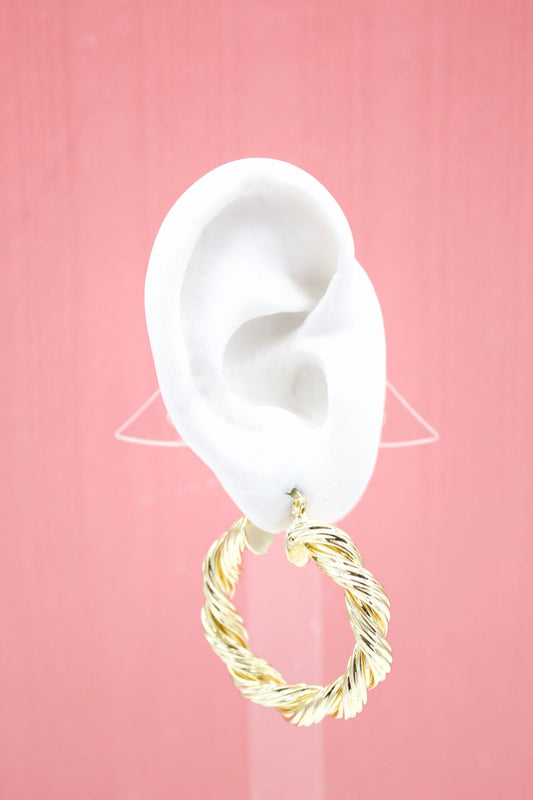 A white mannequin ear displays a chunky, twisted gold hoop earring against a solid pink background.