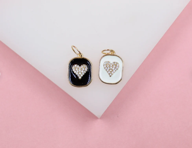 Two rectangular gold-rimmed enamel charms featuring sparkling crystal hearts in black and white sit side by side on a pink and white surface.
