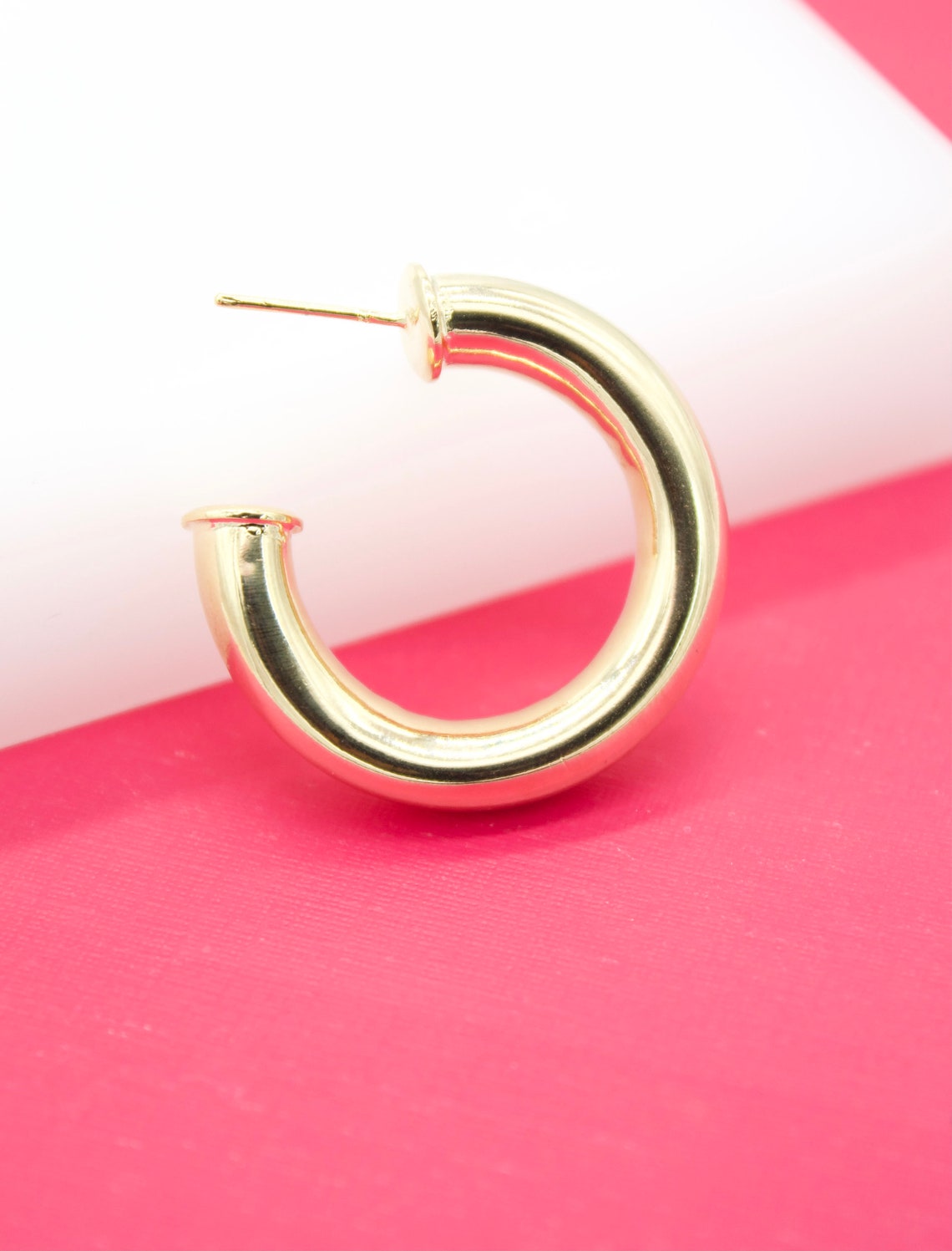 A chunky gold hoop earring rests on a vibrant pink and white background.
