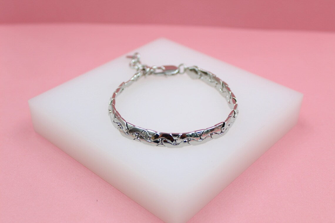 A sleek silver link bracelet rests on a white display block against a soft pink background.