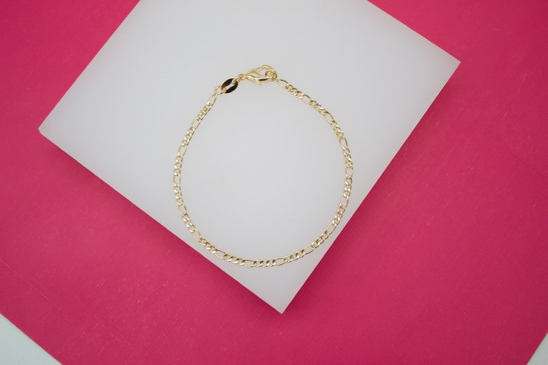 A delicate gold Figaro link chain bracelet rests on a white display surface against a vibrant pink background.