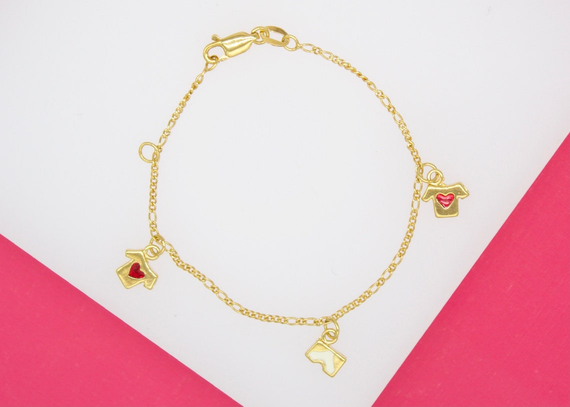 A delicate gold-plated charm bracelet displays miniature t-shirt charms accented with red and white enamel on a white and pink background.
