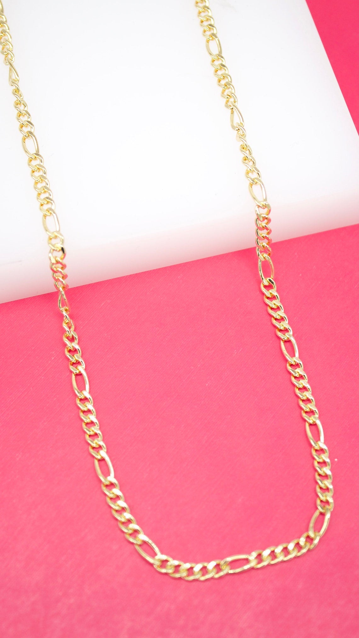 A glossy gold Figaro chain necklace rests against a vibrant pink and white background.