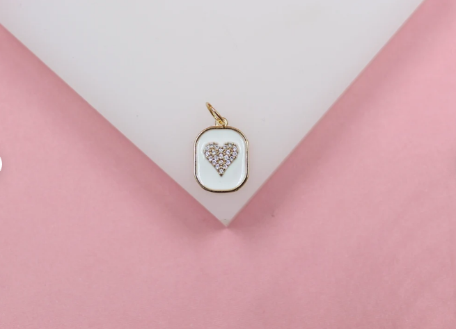 A gold rectangular pendant featuring white enamel and a sparkling pavé crystal heart rests on a white display against a pink background.