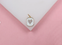 A gold rectangular pendant featuring white enamel and a sparkling pavé crystal heart rests on a white display against a pink background.