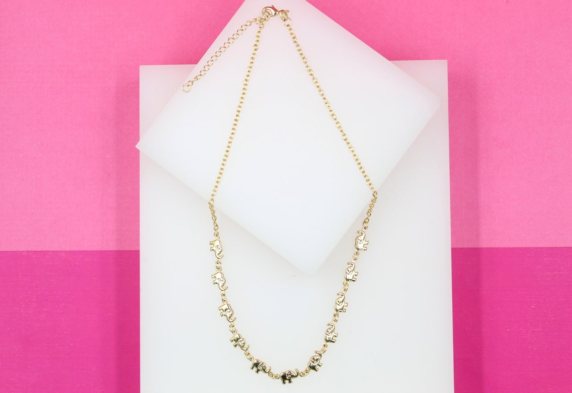 A delicate gold chain necklace featuring miniature elephant charms is displayed on a white platform against a pink background.
