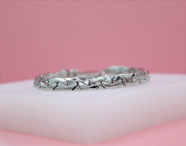 A sleek silver link bracelet rests neatly on a white display block against a soft pink background.