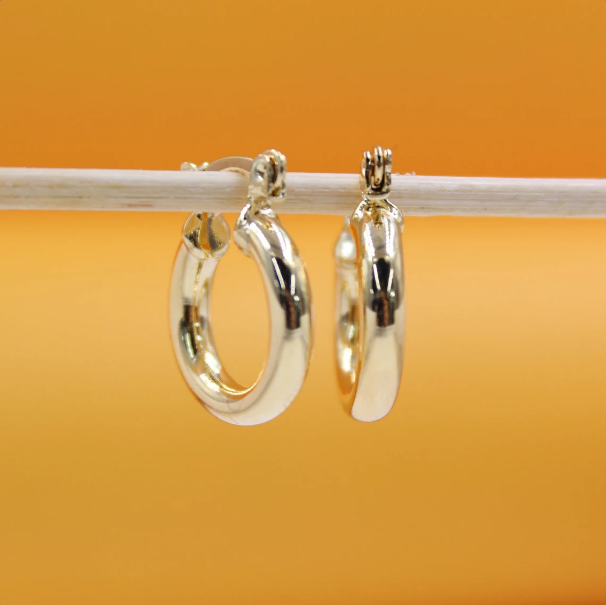 Two polished gold hoop earrings hang side-by-side on a light-colored wooden stick against a warm orange background.