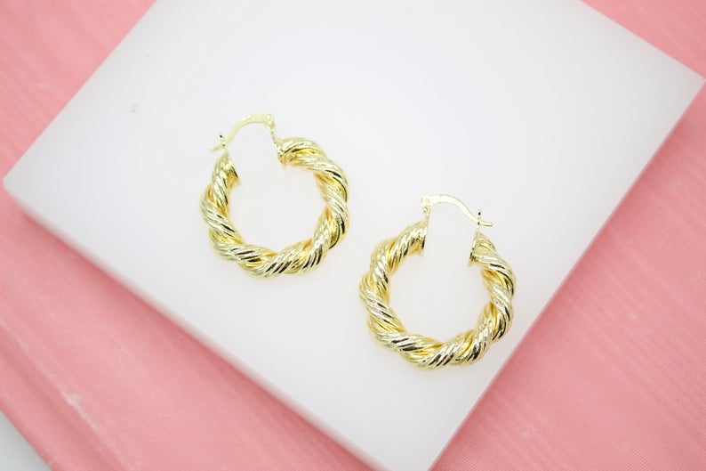 A pair of shiny gold twisted hoop earrings rest on a white display stand against a pink background.