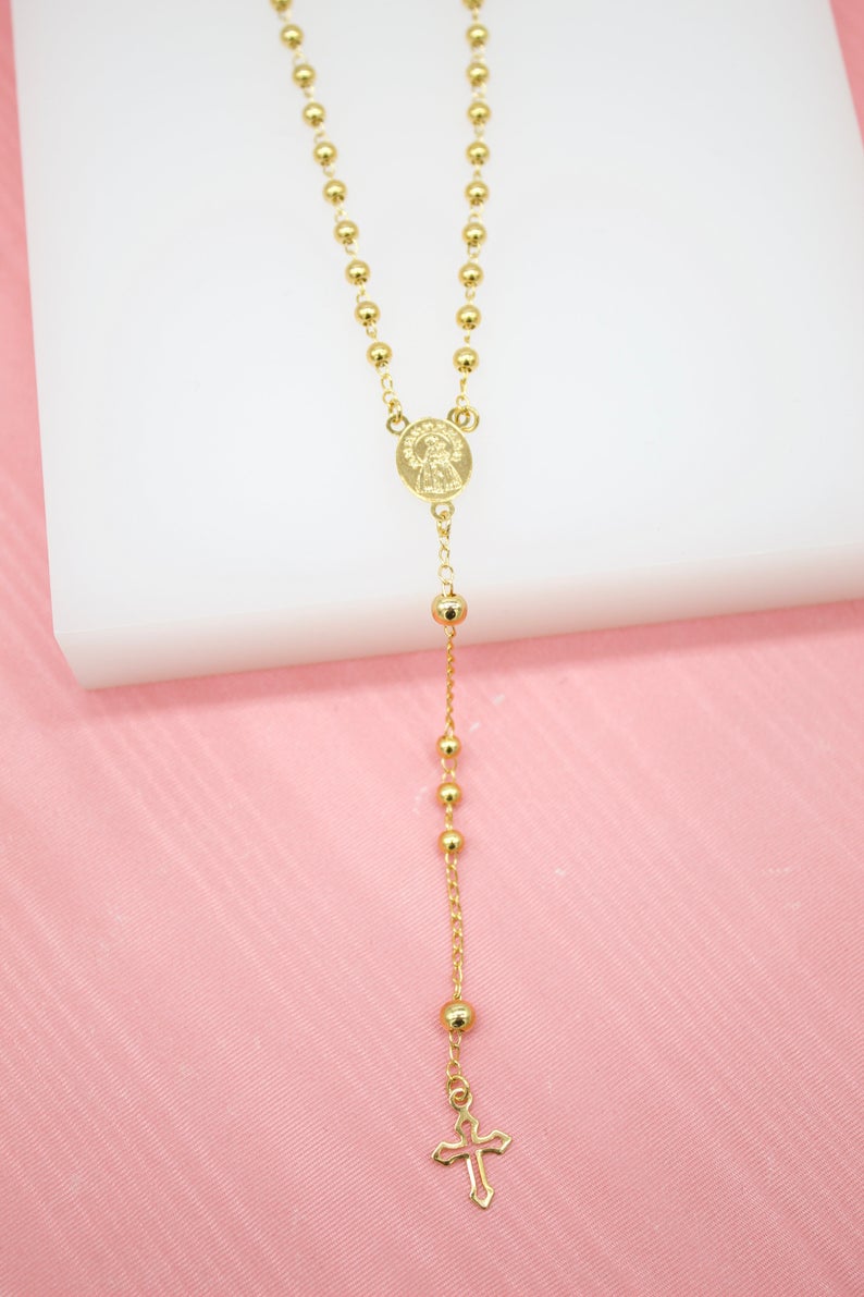 A delicate gold rosary necklace rests against a white and pink backdrop, displaying polished spherical beads, a central medallion, and a dangling cross pendant.