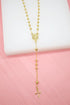 A delicate gold rosary necklace rests against a white and pink backdrop, displaying polished spherical beads, a central medallion, and a dangling cross pendant.