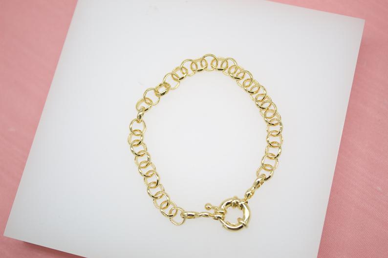 A gold-plated link bracelet rests on a white display surface against a pink background.