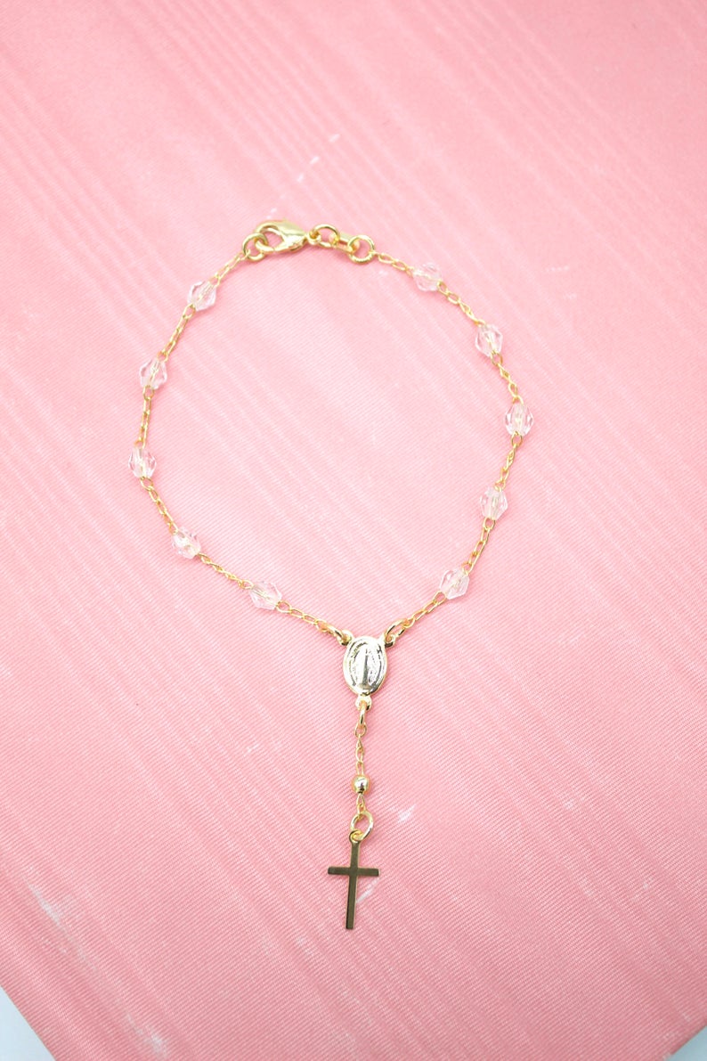 A delicate gold rosary bracelet with crystal beads rests against a vibrant pink background.