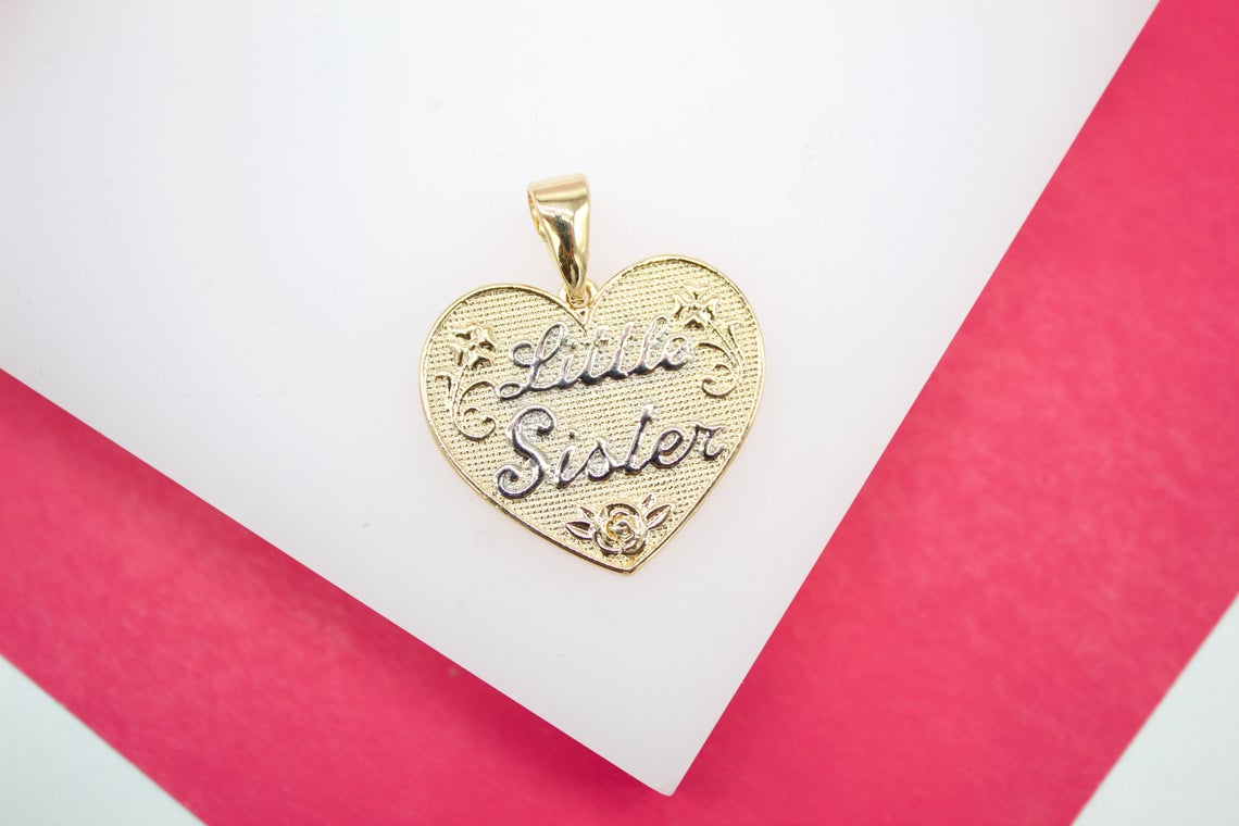 A gold heart-shaped pendant engraved with the words "Little Sister" rests on a white and pink surface.