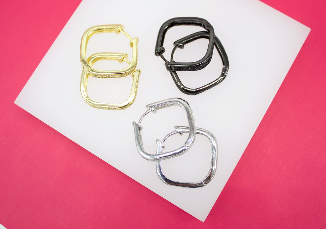 Three pairs of geometric square hoop earrings in gold, silver, and black rest on a white surface against a bright pink background.
