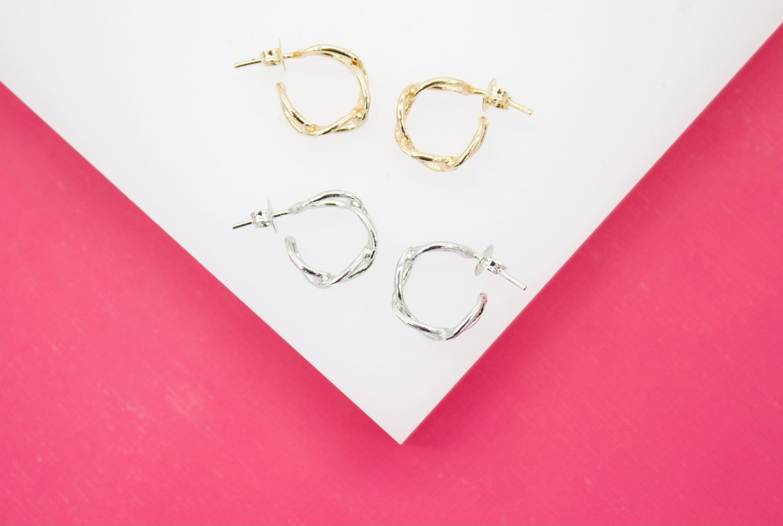 Two pairs of twisted metal hoop earrings in gold and silver rest on a white display surface against a pink background.