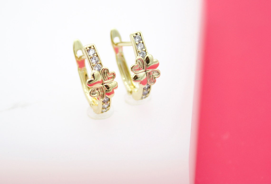 A pair of gold huggie hoop earrings featuring sparkling cubic zirconia stones and a charming pink clover design sits on a white display surface.