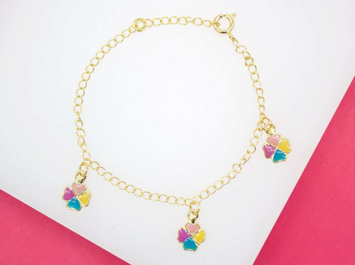 A delicate gold-plated children's chain bracelet features three colorful enamel clover charms displayed on a white and pink surface.