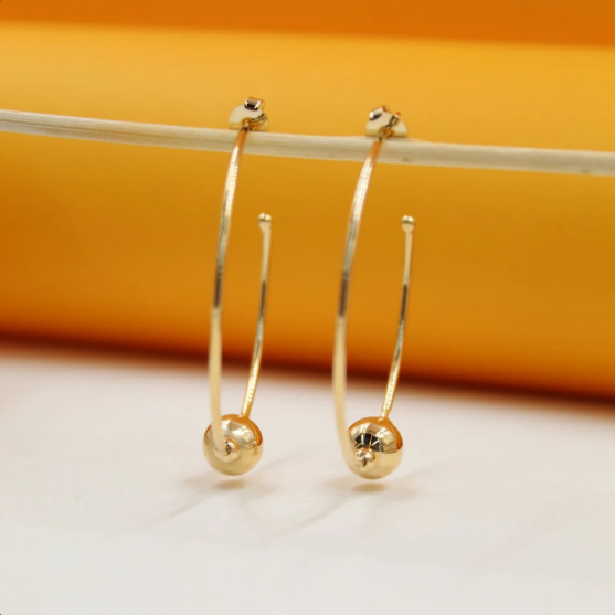 Two elegant gold hoop earrings featuring polished ball charms dangle from a thin horizontal rod against a warm yellow and white backdrop.