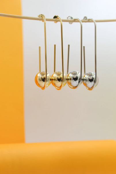 Four dangling Saturn-shaped earrings in gold and silver hang from a thin string against a dual-toned orange and white backdrop.