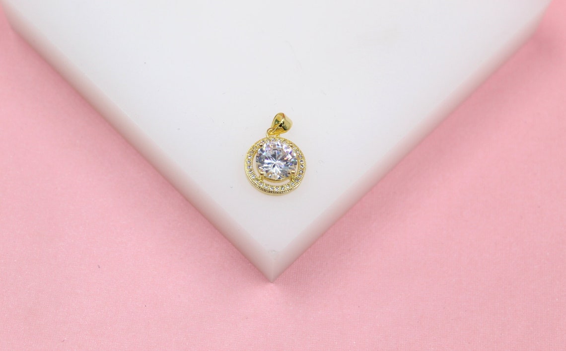 A gold pendant featuring a sparkling round cubic zirconia stone surrounded by a halo of small diamonds rests on a white display surface against a pink background.