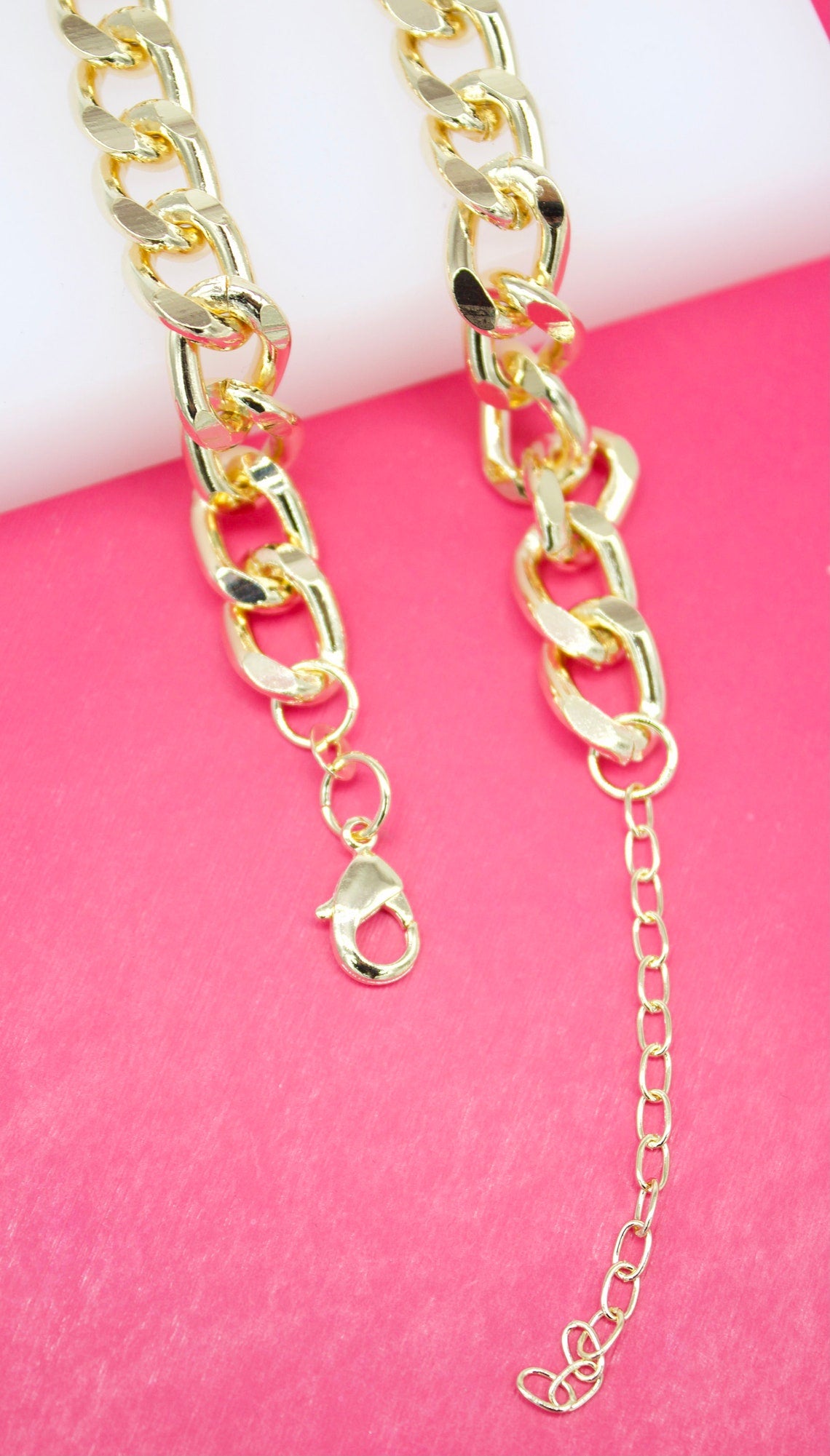 A close-up shot displays a glossy gold-plated curb chain necklace resting against a vibrant pink and white background, highlighting its lobster clasp and adjustable extension chain.