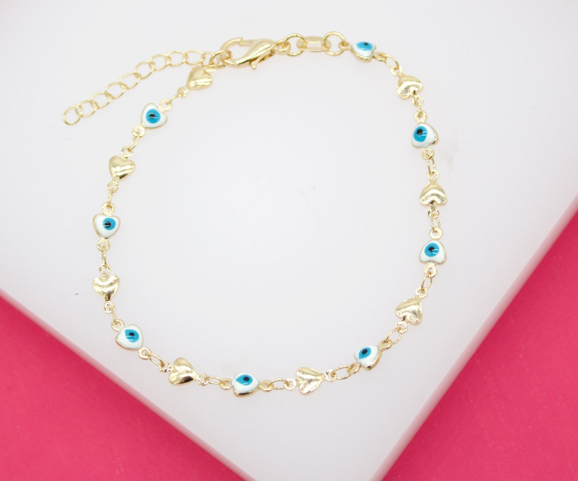A gold chain bracelet features alternating evil eye charms and tiny hearts against a white and pink background.