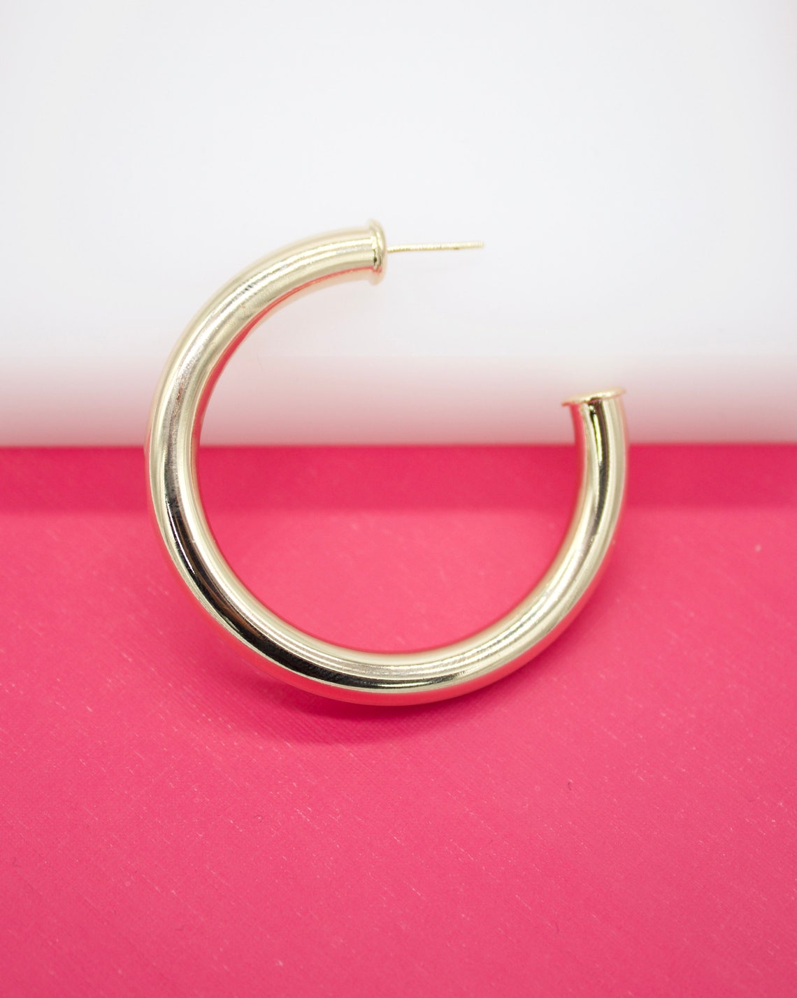 A thick, polished gold hoop earring rests against a vibrant pink and white background.
